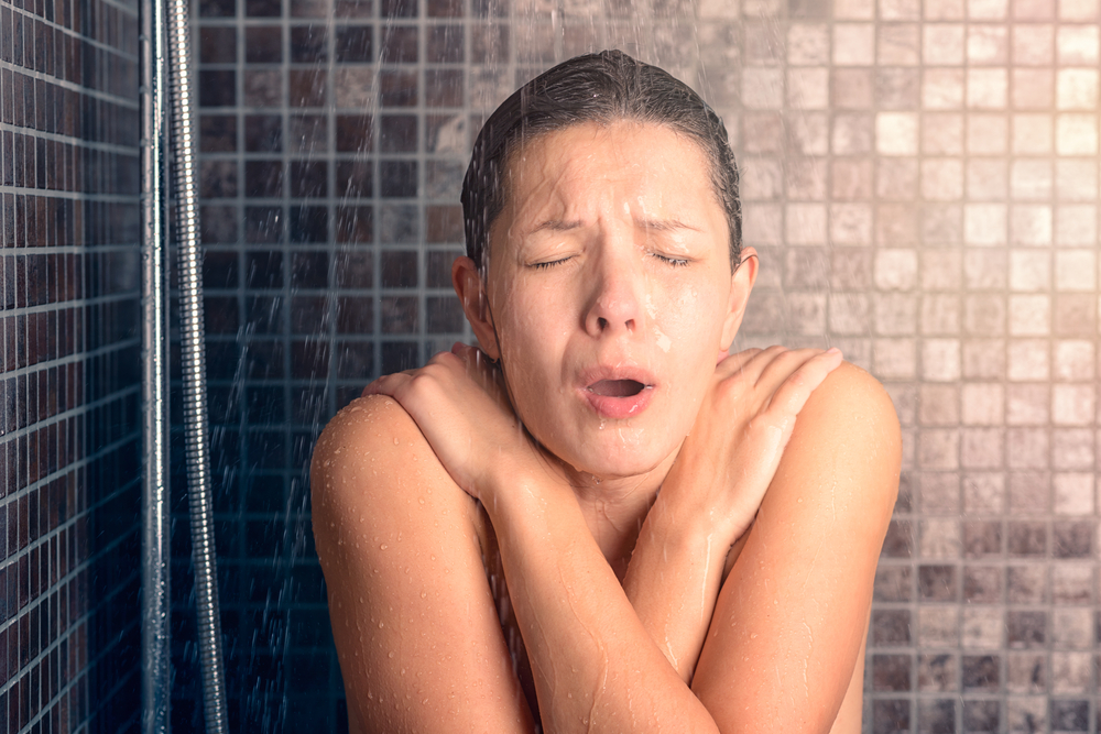 Young woman reacting in shock to hot or cold shower water as she stands under the shower