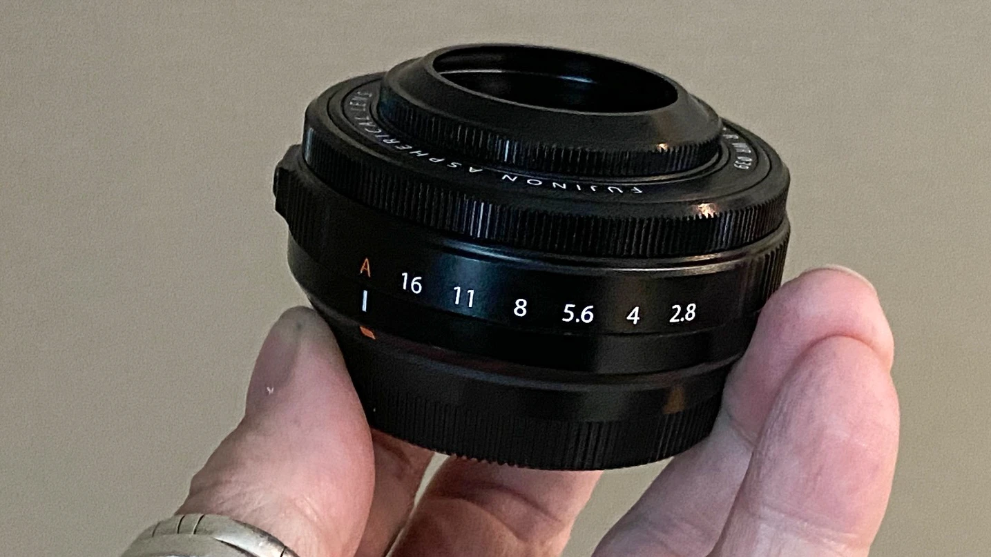 Компактний об'єктив-млинець Fujinon XF 27mm f/2.8 R WR зі шкалою діафрагми у пальцях руки на світлому фоні